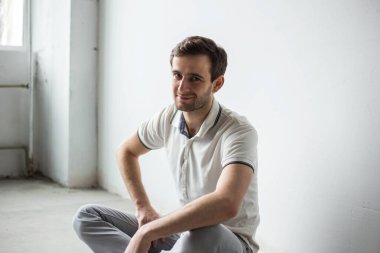 Beyaz tişörtlü ve gri pantolonlu bir adam beyaz bir duvara karşı oturuyor, tek renkli.