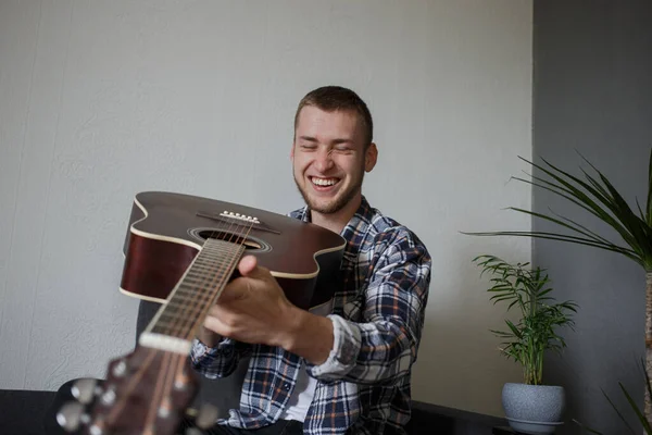 A man in a plaid shirt holds a guitar and laughs