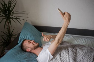 Adam yatağa uzanıyor ve baş parmağıyla selfie çekiyor..