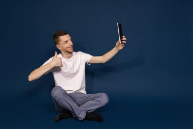 Beyaz tişörtlü, mavi arka planda oturan bir adam tabletten selfie çekiyor.