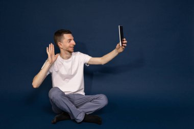 Beyaz tişörtlü, mavi arka planda oturan bir adam tabletten selfie çekiyor.