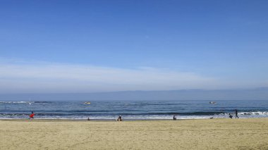 Sahilde sabah, bir grup insan deniz kıyısında rahatlarken, sörfçüler, evcil hayvanlarıyla gezerken ya da oynarken eğleniyor..