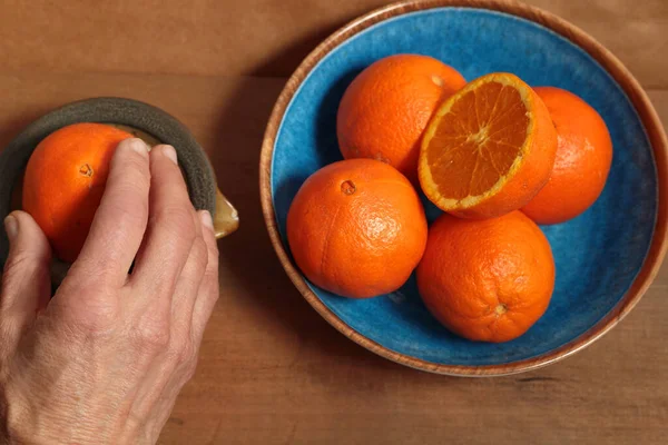 Hand squeezing an orange to prepare juice, next to it blue plate with some fruits