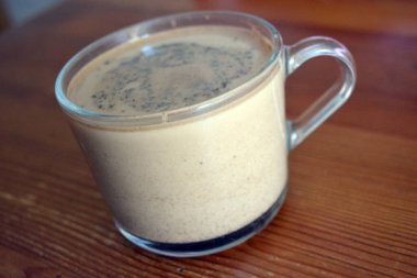 A transparent pleasant glass cup with natural brewed coffee with milk is located on a dark yellow wooden table.