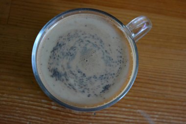 A transparent pleasant glass cup with natural brewed coffee with milk is located on a dark yellow wooden table.