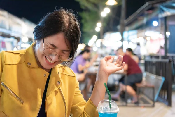 Güzel Asyalı kadın (LGBTQ) doğal makyaj, Tayland sokak yemekleri, market veya restoran modasında yiyecek veya içecek ile poz veren gözlüklü sarı deri kıyafetler giyer.