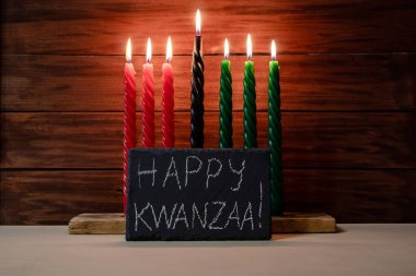 Happy Kwanzaa. African American holiday. Seven candles red, black and green on wooden background. Symbols of African heritage. Congratulatory inscription on chalk board. clipart