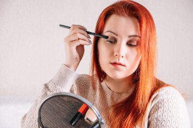 Kızıl saçlı genç kadın makyaj yapıyor. Bayan aynaya bakar ve kozmetik kullanır..