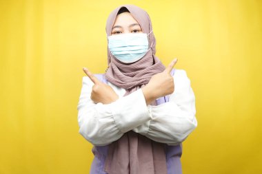 Tıbbi maske takan Müslüman bir kadın, eli boş bir yeri işaret ediyor, eli yukarıda sarı arka planda izole edilmiş bir şeyi gösteriyor.