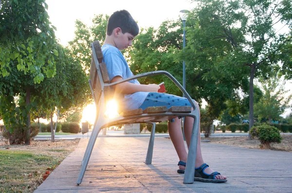 İspanyol Çocuk parkta arka planda gün batımı olan bir bankta mobil eğlence seti oynuyor.