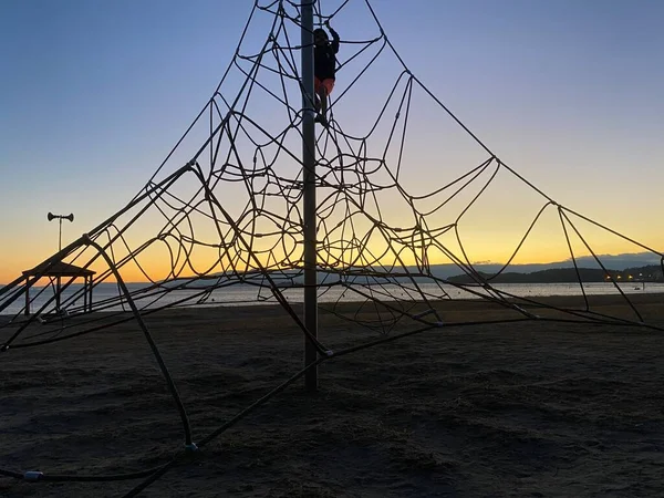 Silhouette of a child climbing in a rope playground structure on a beach during sunset