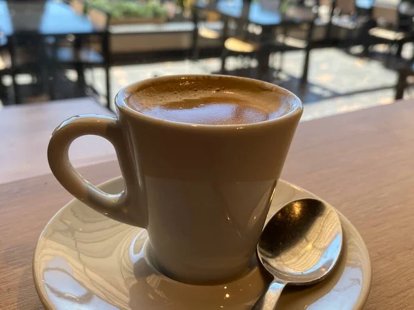 Close up of espresso coffee. Sunlight over the delicious foam in the top. White plate , mug and a spoon below. Wooden table and nice terrace background. Relax and take a break concept
