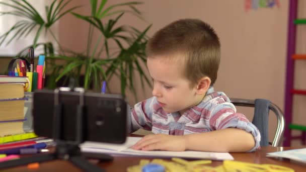 El niño aprende de forma remota con el teléfono inteligente. El niño pequeño se sienta a la mesa. Aprendizaje a distancia durante cuarentena. — Vídeo de stock