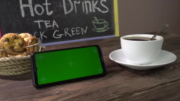 Varm drikke, cookies og smarttelefon på bordet på kafeen. Smartphone green screen. – stockvideo