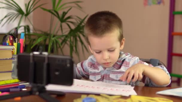 Хлопчик дошкільного віку показує літери, написані на аркуші, дивлячись на смартфон. . — стокове відео