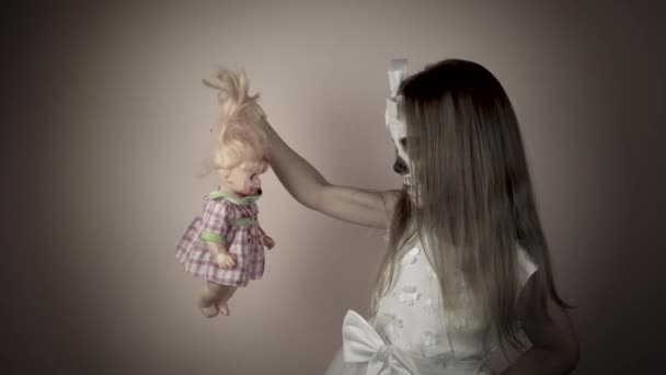 Маленькая девочка в белом платье с раскрашенным лицом держит окровавленную куклу в руке и смотрит на нее.. — стоковое видео