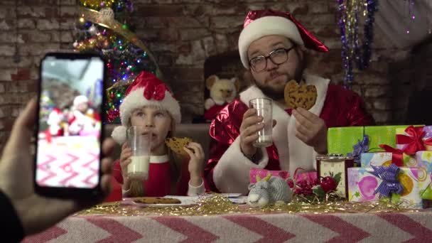 Um homem vestido de Papai Noel e uma menina estão bebendo leite e cookies.Posing para smartphone. — Vídeo de Stock