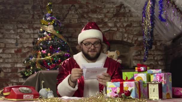 Père Noël à la maison est assis à une table avec des cadeaux et passe à travers des enveloppes avec des lettres au Père Noël. — Video