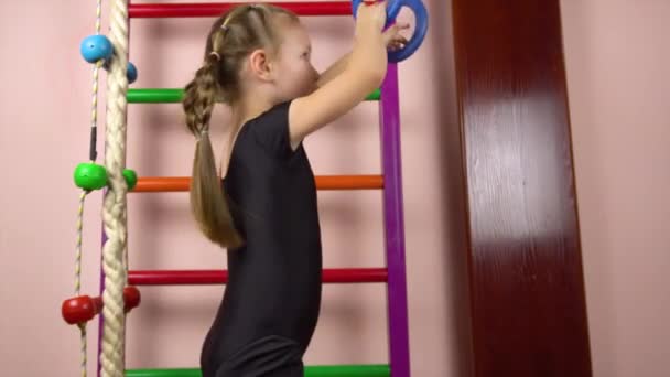 Ένα μικρό όμορφο κορίτσι πέντε ετών πηγαίνει για σπορ σε γυμναστικούς δακτυλίους.Σουηδικό τοίχο. — Αρχείο Βίντεο