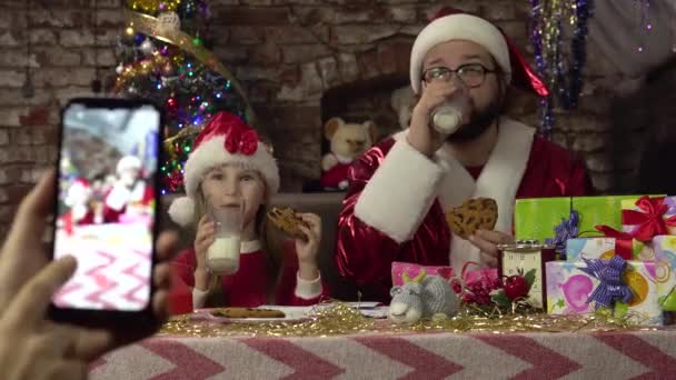 Чоловік, одягнений як Санта і маленька дівчинка п'ють молоко і печиво. Поза смартфоном.. — стокове відео