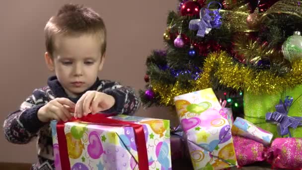 Little curious boy sitting under the Christmas tree unties the ribbon on the gift box.Christmas. — Stock Video