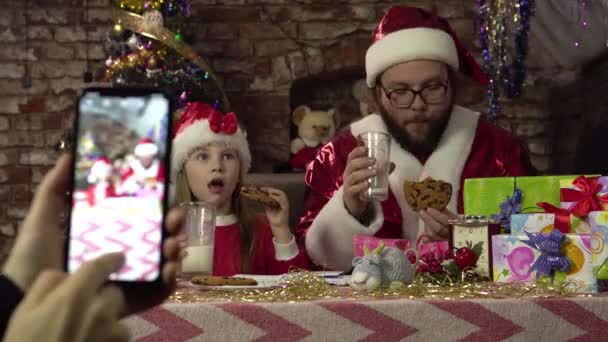Muž oblečený jako Santa a holčička pijí mléko a sušenky.Pózování pro smartphone. — Stock video