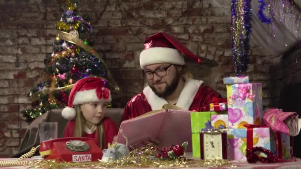 Маленькая девочка и Санта-Клаус сидят за столом с подарками и читают книги.4k — стоковое видео