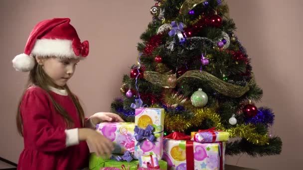 Een klein meisje in een rode jurk en een kerstman hoed is het kiezen van een doos met een geschenk onder de boom.4k — Stockvideo