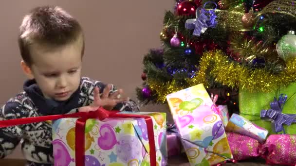 Menino curioso sentado sob a árvore de Natal desata a fita na caixa de presente.Natal. — Vídeo de Stock