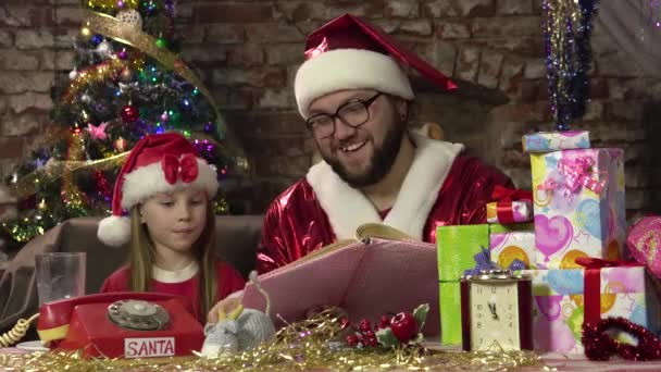 Santa Claus se sienta en el sillón y lee el libro con cuentos de hadas para la niña entusiasta — Vídeos de Stock