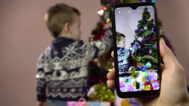 Küçük çocuk Noel ağacını süslüyor ve akıllı telefondan çekim yapmak için poz veriyor.. — Stok video