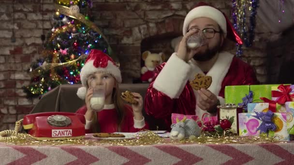 Noel Baba ve küçük bir kız gibi giyinmiş bir adam süt ve kurabiye içiyor. Sosyal ağlar için fotoğraf. — Stok video
