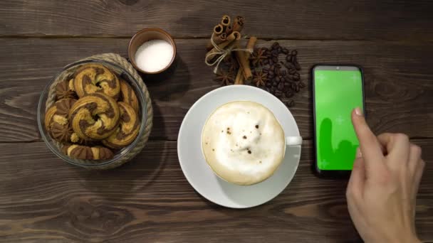 Una mano femenina presiona la pantalla verde del teléfono inteligente que está sobre la mesa con una taza de café. — Vídeos de Stock