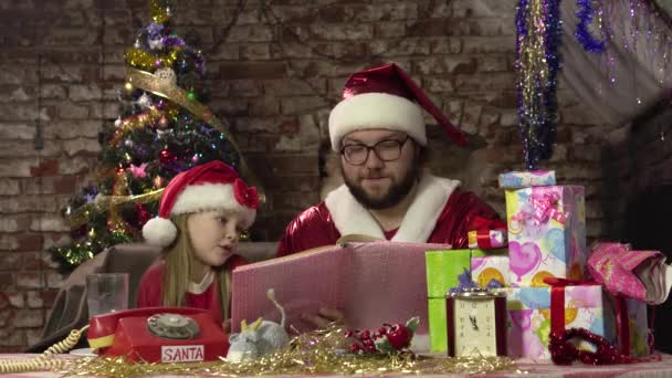 Санта Клаус сидить на кріслі і читає книгу з казками для захопленої маленької дівчинки.. — стокове відео
