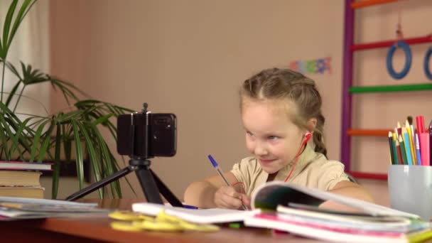 Petite écolière écrit avec un stylo dans un carnet, à une table avec un smartphone. Enseignement à distance. — Video