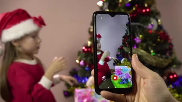 Ein kleines Mädchen als Weihnachtsmann verkleidet schmückt einen Weihnachtsbaum und posiert für Shootings mit dem Smartphone. — Stockvideo