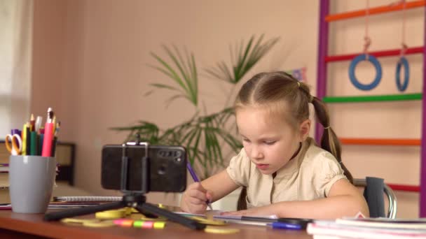 Kleine Schulmädchen schreiben mit dem Stift in ein Notizbuch, an einen Tisch mit dem Smartphone. Fernstudium.