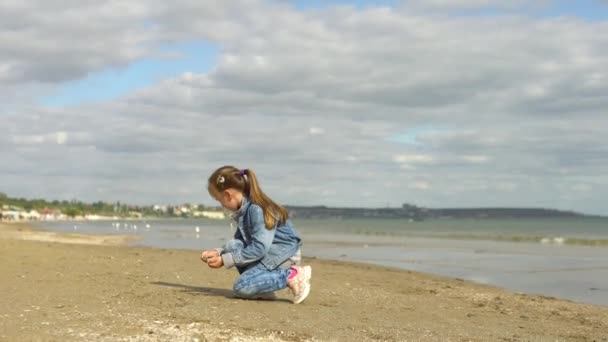 Dziewczynka w dżinsowym garniturze wrzuca muszelki i kamyki do morza. Jesienią jest nad morzem. — Wideo stockowe