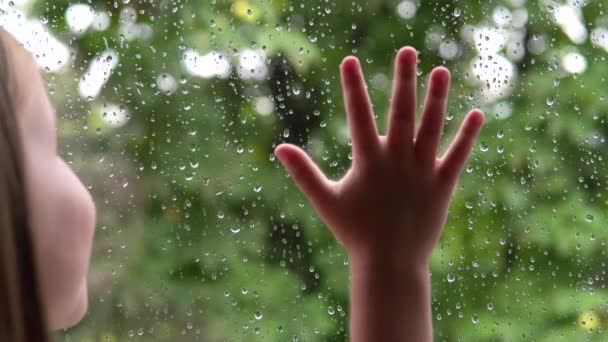 Маленька дівчинка дивиться у вікно під дощем і зеленими деревами. Рука на склі . — стокове відео