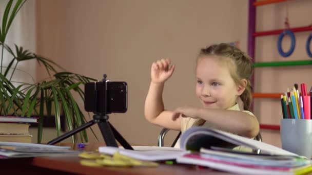 Маленькая школьница сидит за столом со смартфоном и поднимает руку, чтобы ответить. — стоковое видео