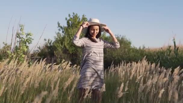 Hermosa chica joven en un sombrero de paja. Ella se para en un campo y mira el sol poniente. — Vídeo de stock