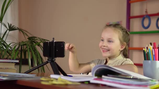 スマホを持ってテーブルに座っている少女が手を挙げて答えます. — ストック動画