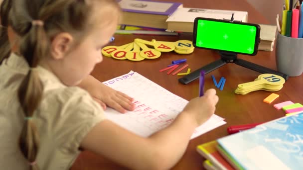 Kleine Schülerin schreibt lateinische Buchstaben am Tisch sitzend mit einem Smartphone mit grünem Bildschirm.4k — Stockvideo