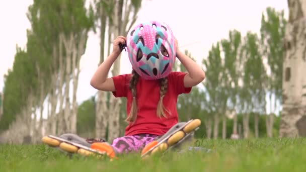 En liten vacker flicka rätar ut en skyddande hjälm medan du sitter på gräset i rullskridskor. — Stockvideo