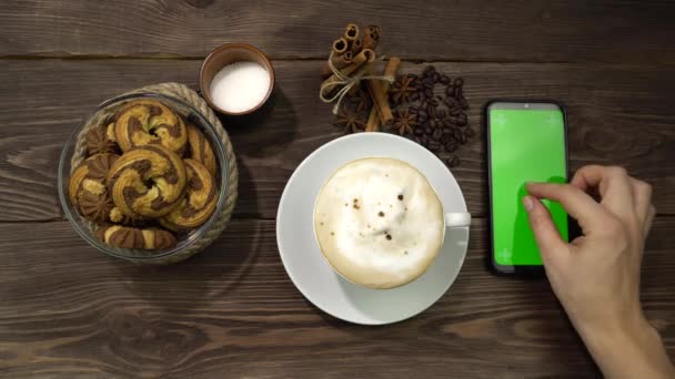 Een vrouwelijke hand drukt op het groene scherm van de smartphone liggend op de tafel met een kopje koffie. — Stockvideo