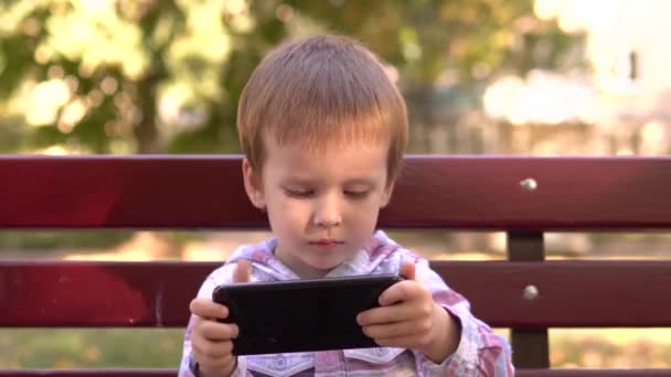 Πορτρέτο ενός μικρού αξιολάτρευτου αγοριού που κάθεται σε ένα παγκάκι και παίζει με ένα smartphone. — Αρχείο Βίντεο