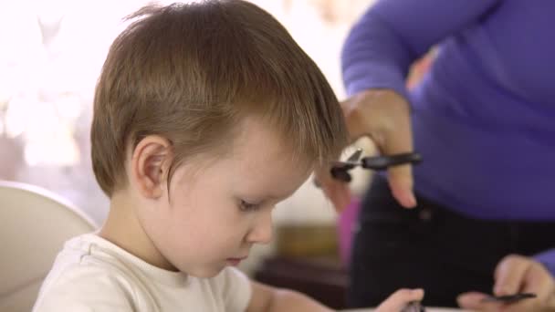 Ένας κομμωτής κουρεύει ένα όμορφο τρίχρονο αγόρι στο σπίτι ενώ παίζει με το smartphone του. — Αρχείο Βίντεο