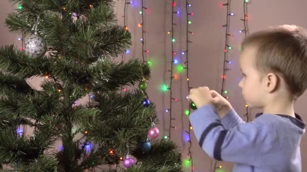 De jongen hangt ballen aan de kerstboom tegen de achtergrond van knipperende bloemenslingers. — Stockvideo