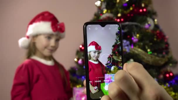 Een klein meisje verkleed als kerstman versiert een kerstboom en poseert voor het schieten op een smartphone. — Stockvideo