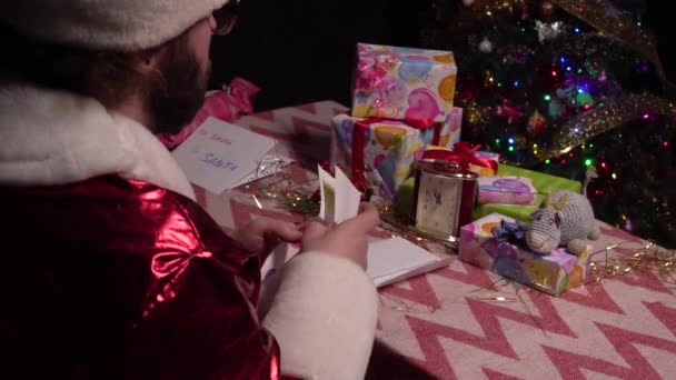 Père Noël, assis à une table avec des cadeaux, déploie une lettre d'enfants demandant d'arrêter Covid 19.4k — Video